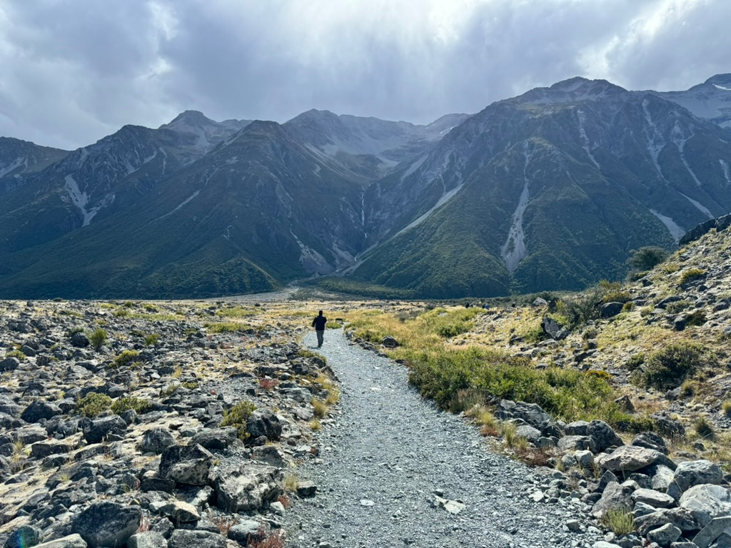 A man walking down a path from Tasman Lake with mountains in the distance in New Zealand