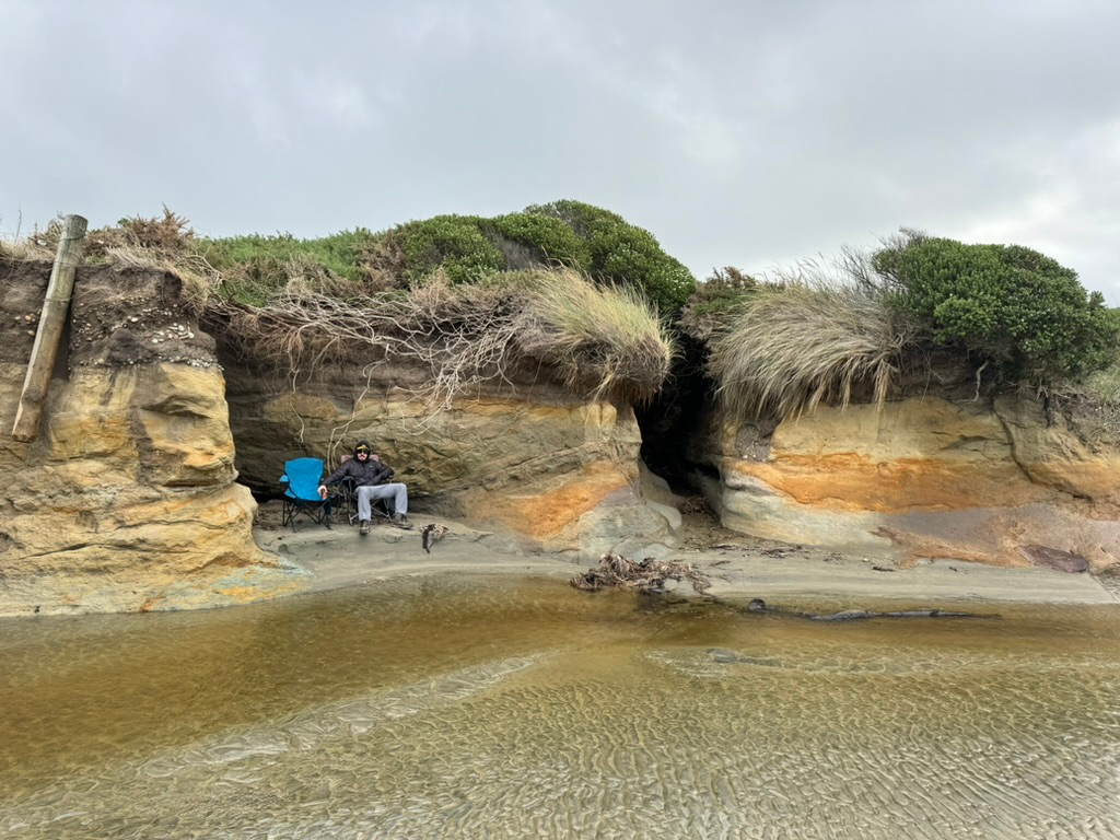 Man in camping chair in front of interesting land formations at Monkey Beach freedom camping in New Zealand