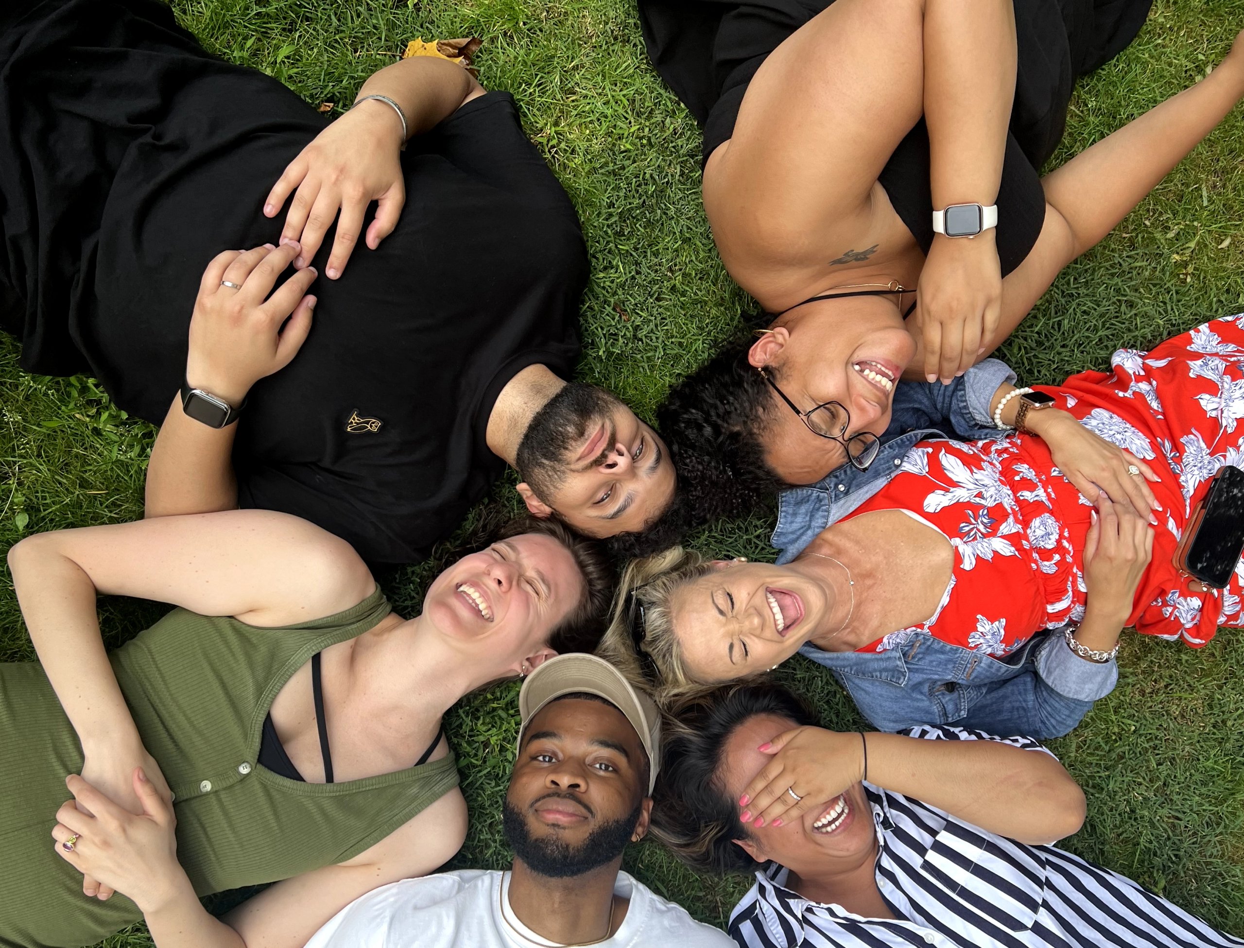 A diverse group of people lying in a circle on the grass with their heads touching in the center, laughing with big smiles