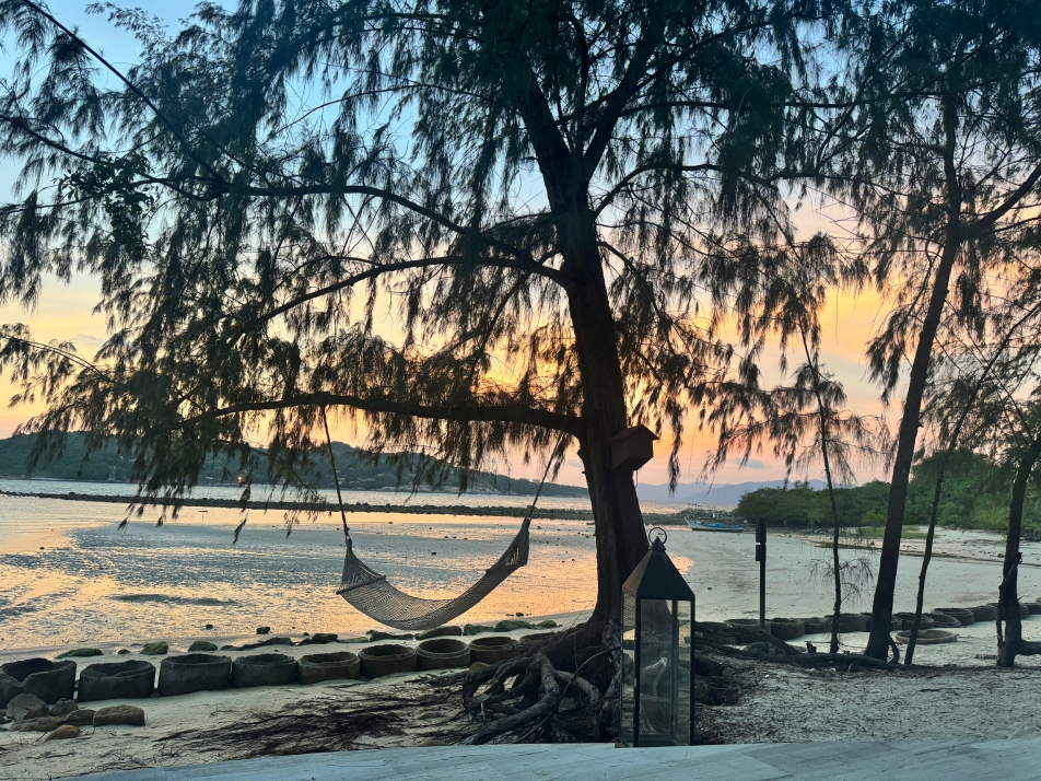 A tropical view of a hammock hanging from a a tree in front of the ocean and a sunset from Cape Fahn Resort on Koh Samui, Thailand