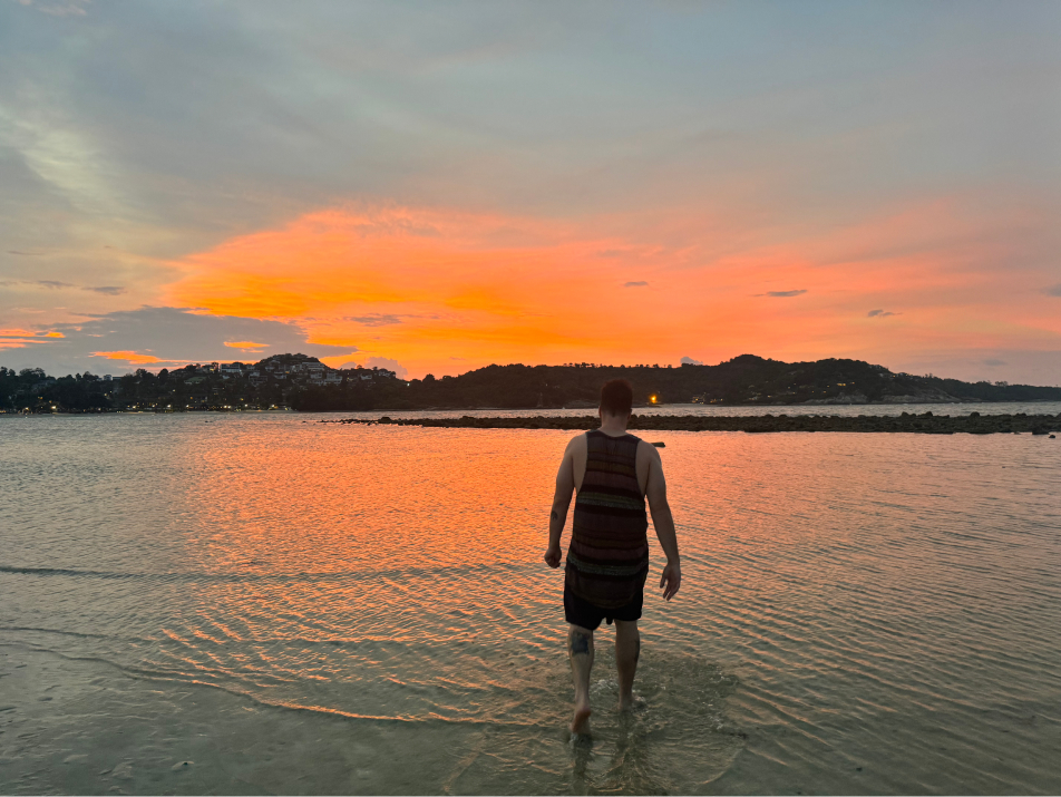 A man walking through shallow ocean water toward a stunning orange sunset on Koh Samui, Thailand