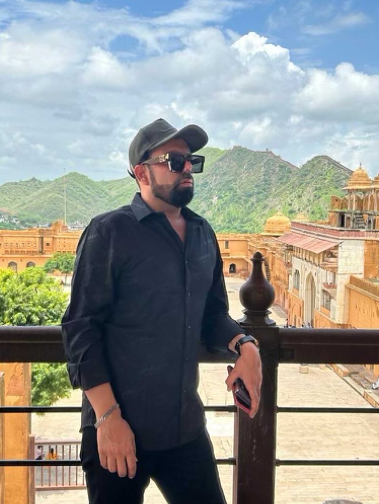 An Indian man with a baseball cap and sunglasses in front of a scenic view in India