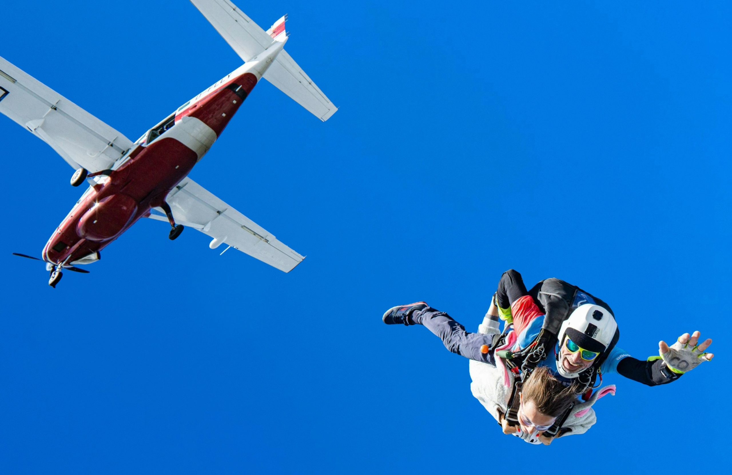 A woman tandem skydiving with a man, with a view of the airplane that they just jumped out of in the background