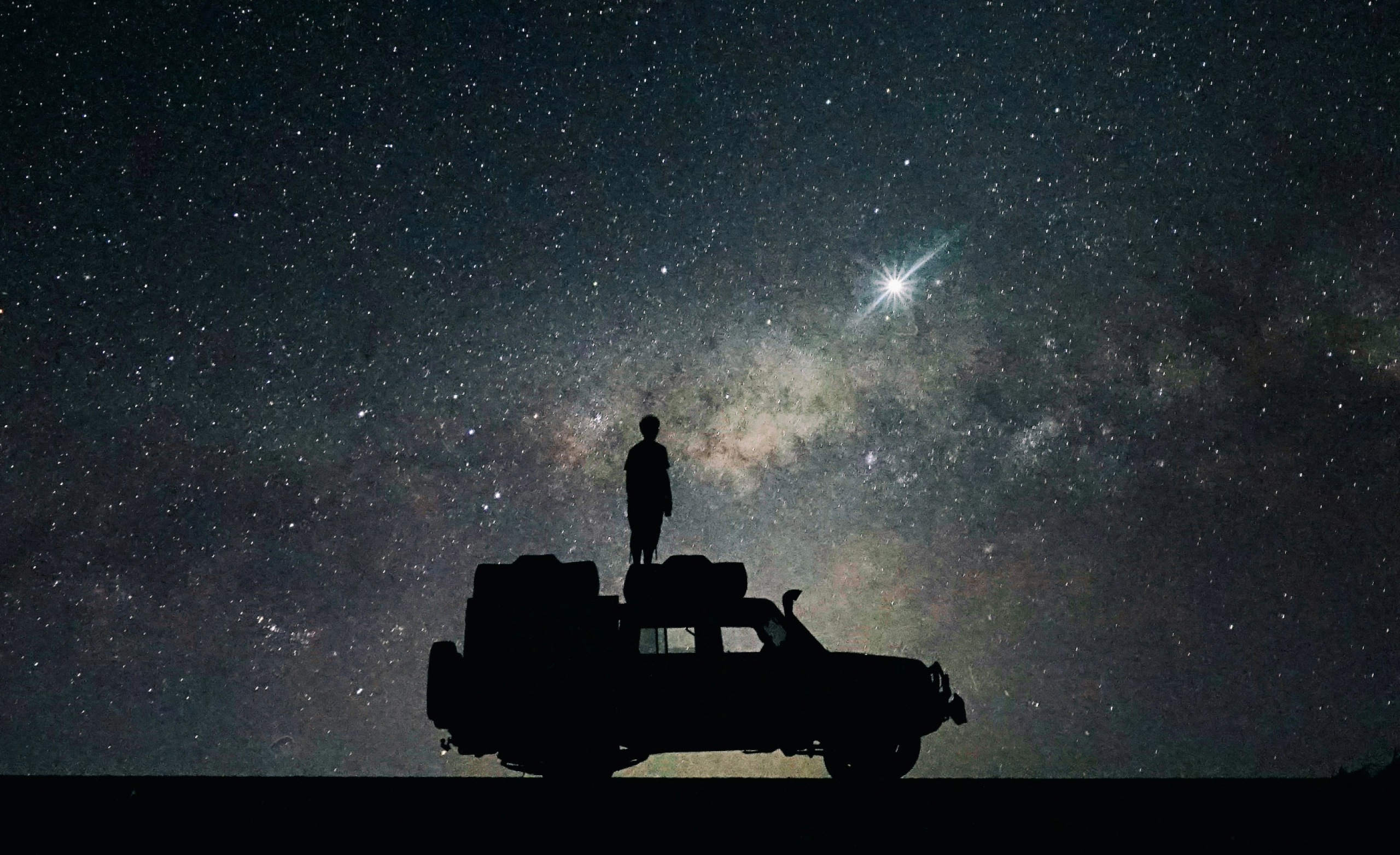 A silhouette of a person standing on top of their camper van / truck looking up at a very starry sky at nighttime