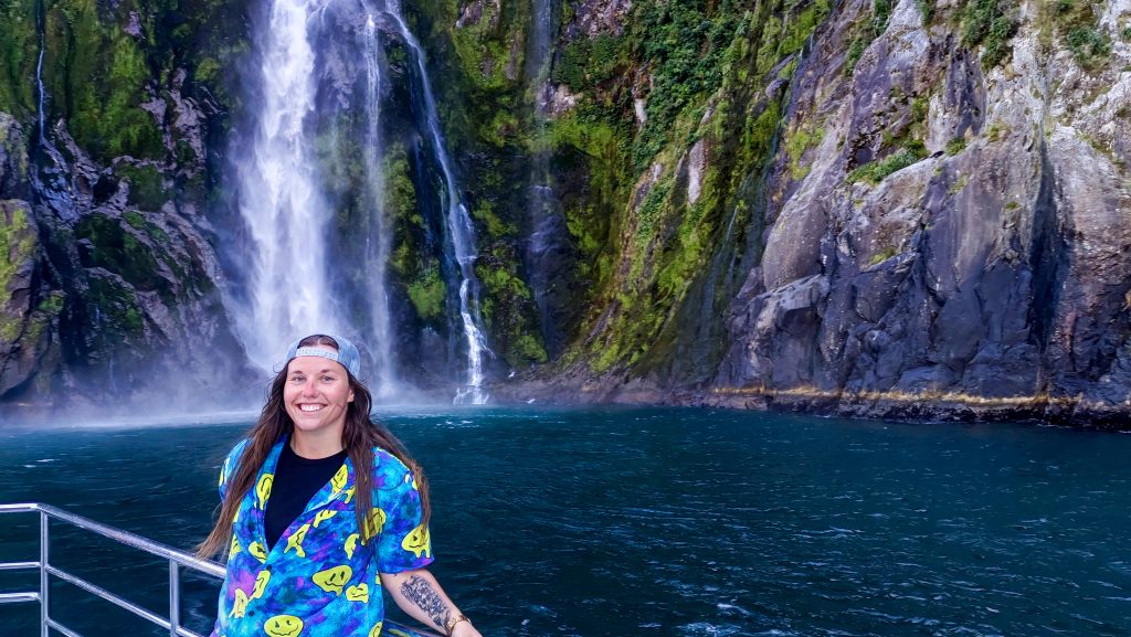 A woman with a fun t-shirt and backwards cap in front of a waterfall in New Zealand