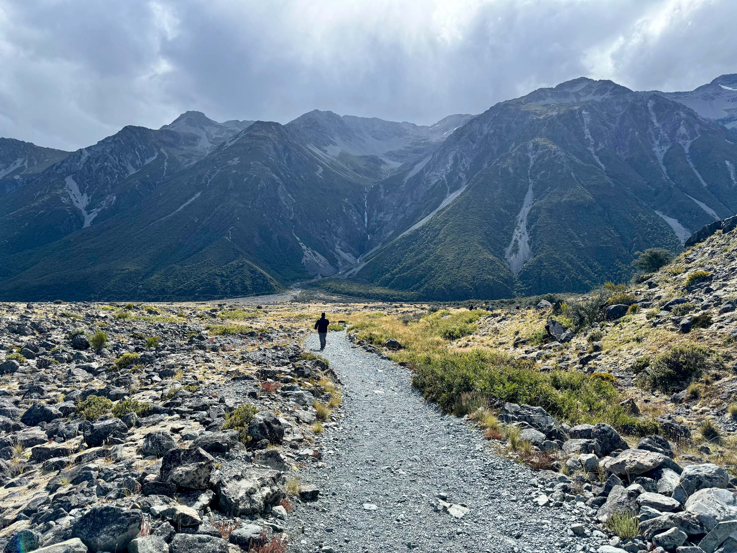 A man walking down a gravel path toward imposing mountains in Aoraki / Mount Cook, New Zealand