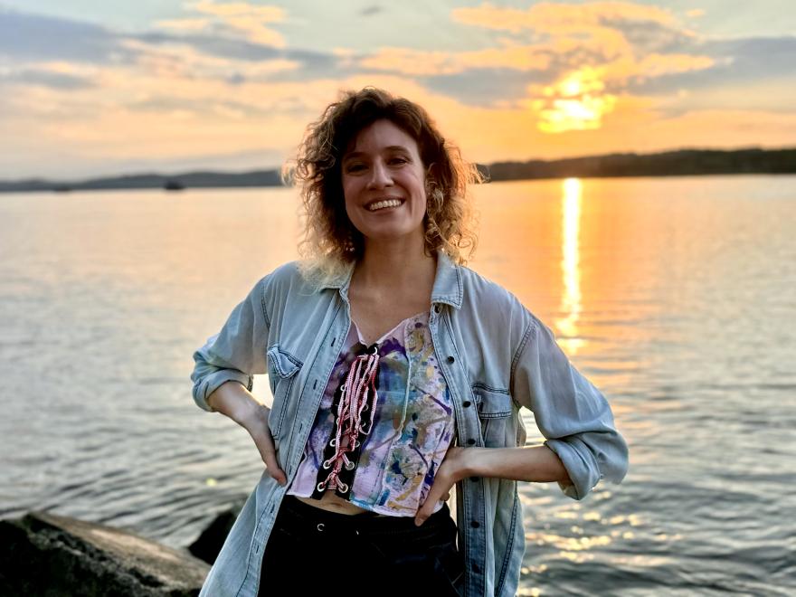 A woman with her hands on her hips and a big smile, dressed casually, standing in front of the Hudson River and a beautiful sunset in New York, USA