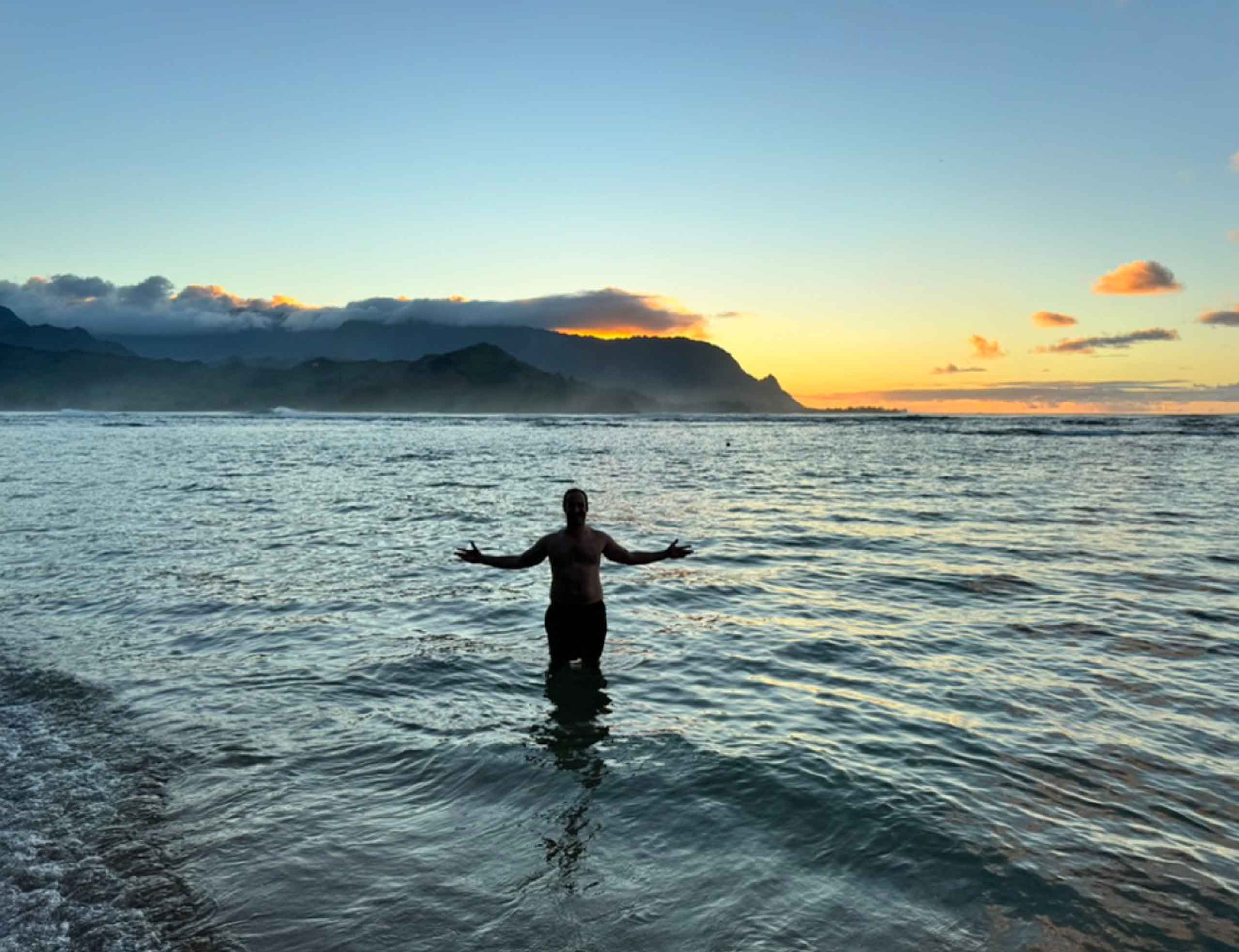 Man wading in water with arms out and sunset in background at 1 Hotel Hanalei Bay in Kauai, Hawaii, USA